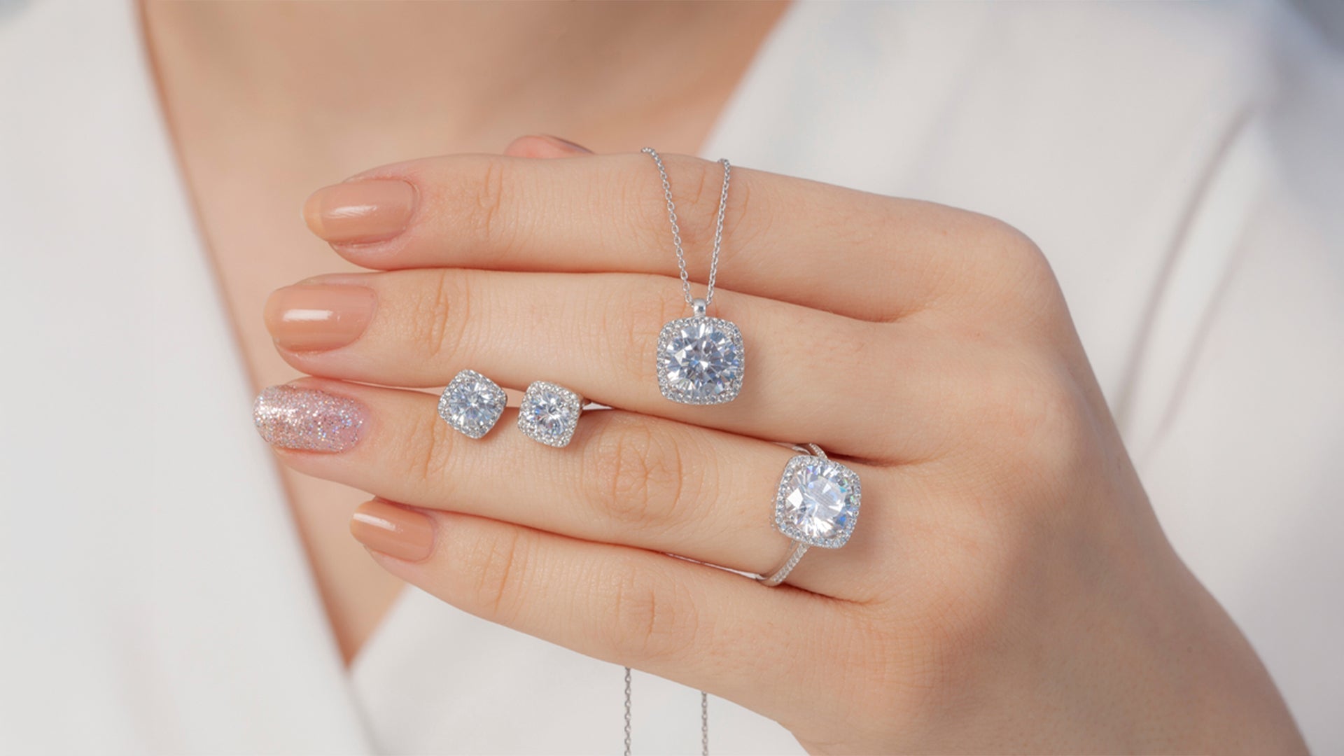 A woman's hand displaying elegant jewelry like diamond stud earrings, a sparkling ring, and a pendant on a delicate chain.