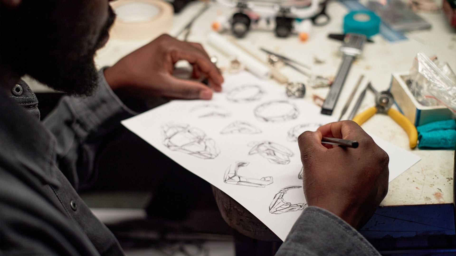 A person sketches ring designs on paper at a cluttered workbench, surrounded by tools and materials, conveying creativity and focus in a jewelry-making setting.