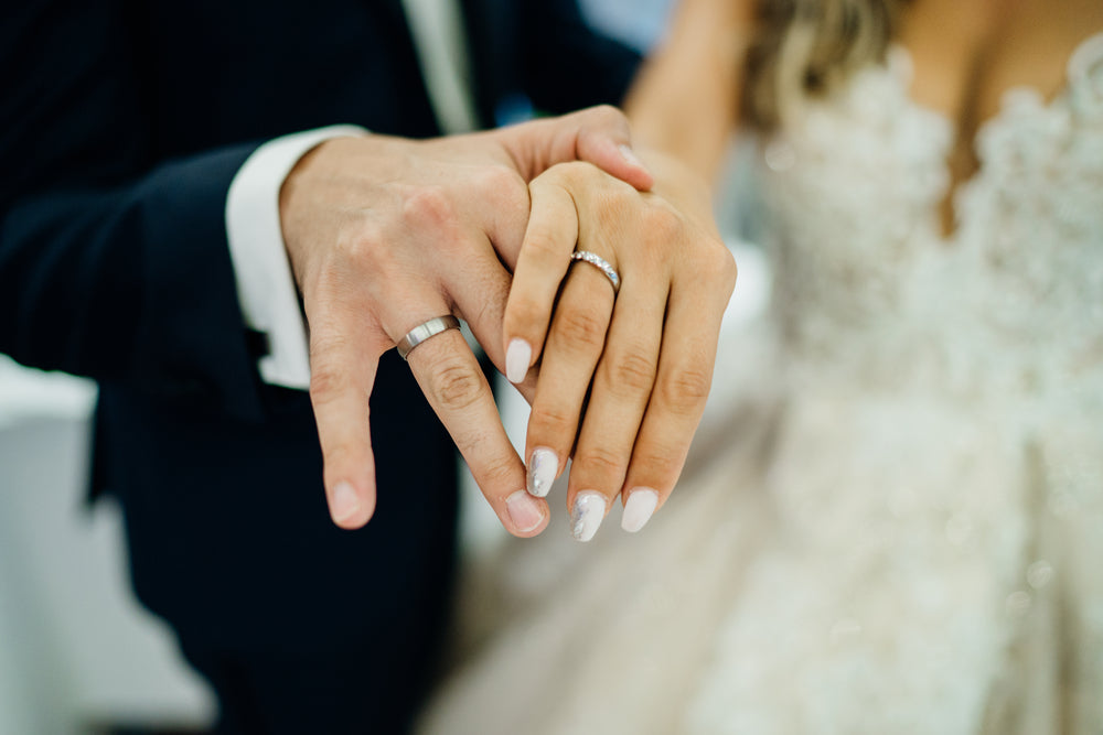 Close-up of a couple's hands, both wearing wedding rings. The background shows a blurred wedding dress and suit, conveying a joyful, romantic moment.