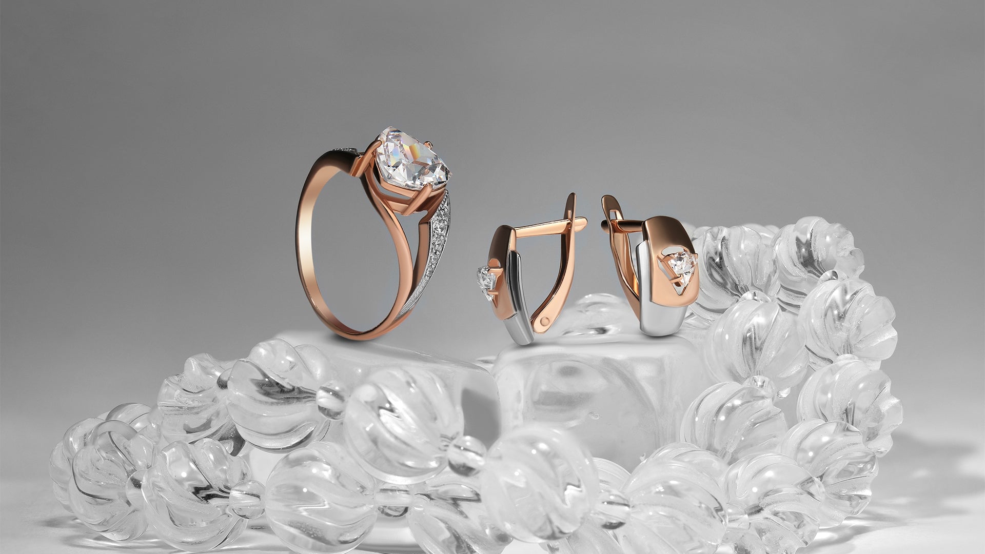 A set of rose gold earrings and a ring studded with diamond. They are sitting on a clear, sculpted, crystal-like surface